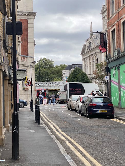 A narrow urban street scene during daytime showing parked cars lining the curb on the right side, with a black van and a white box truck positioned near a building entrance on the right, part of a home relocation or removals process. The street has double yellow lines along the edge indicating parking restrictions and is bordered by multi-storey brick and stone buildings, some with signage and window displays. In the background, a temporary pedestrian crossing with barriers and a group of people are visible near a storefront decorated with banners. The overcast sky above contributes to diffuse lighting, typical of a busy city environment where furniture transport and packing activities might be taking place as part of a house moves service. The scene includes elements such as plastic wrapping, cardboard boxes, and lifting equipment associated with professional removals and moving logistics, while [COMPANY_NAME]'s presence is implied through the context of service provision.