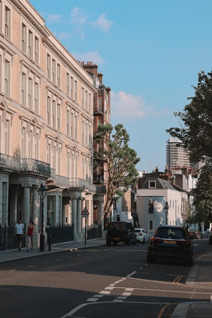 A street scene showing a building with a modern glass and concrete facade on the left and a traditional stone building with rounded windows and decorative elements on the right, with a large tree in the center partially obscuring the view. In the foreground, there is a paved area with a few pedestrians walking, including one person with a backpack. A moving van with a company logo is parked near the tree, and a worker is loading or unloading boxes and furniture wrapped in plastic and blankets from the van. The scene suggests a home relocation process involving furniture transport and packing, with members of the Man with Van Bayswater team managing the loading on a cloudy day.