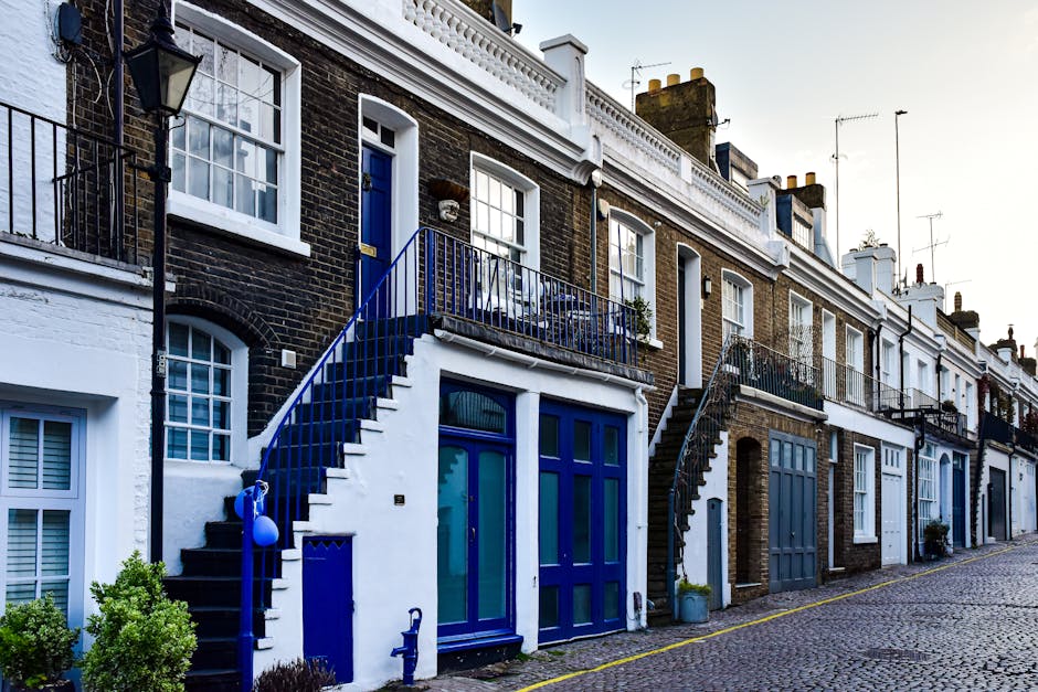 View of a narrow street in Bayswater featuring a vintage beige compact car parked directly in front of a residential building with a white facade and arched doorway. The building has three floors with